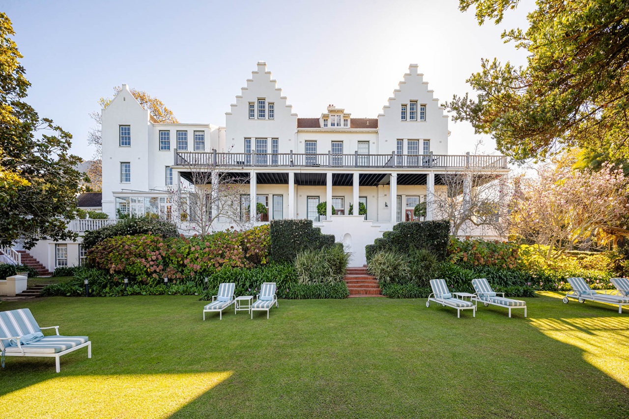 The Cellars-Hohenort Hotel bei Sonnenschein, mit gepflegtem Park und Sonnenliegen auf einer Wiese im Vordergrund.