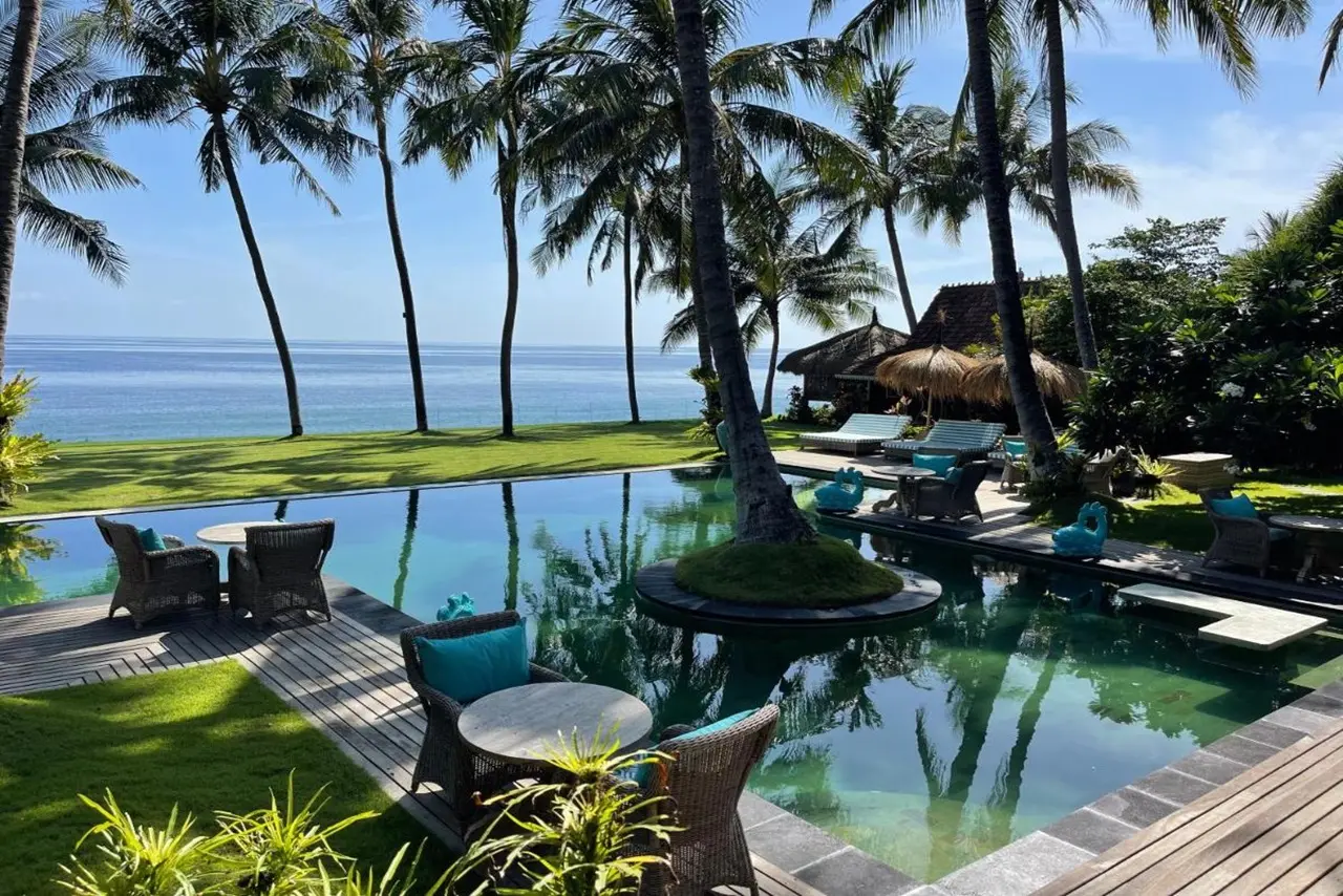 Pool mit Korbmöbeln, Palmen, Strand und Meer im Louka Beach Bali Resort bei Sonnenschein.